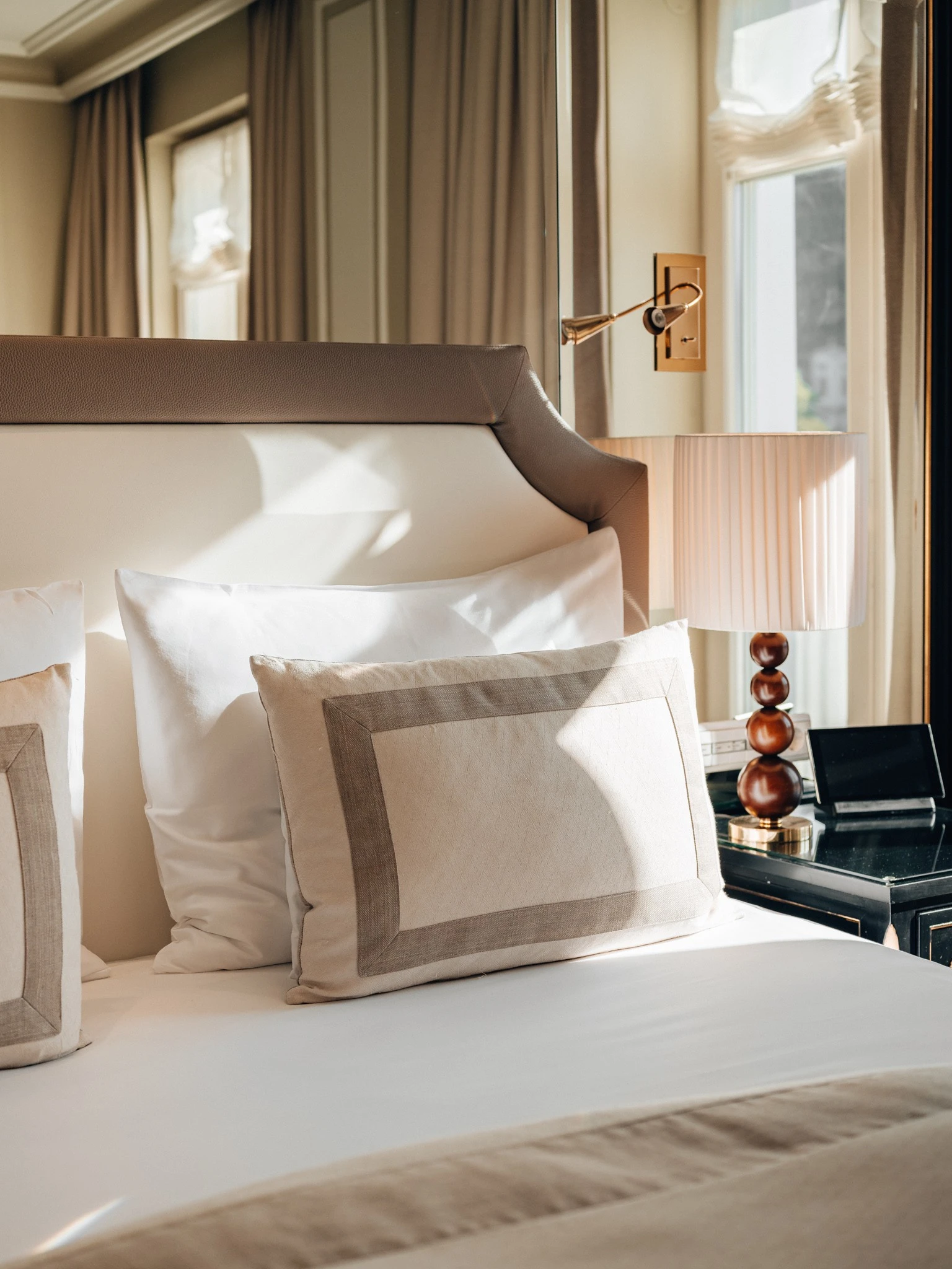 Sunlight gently pours onto a neatly made bed with crisp white linens and beige-accented pillows, beside a stylish nightstand with a sculpted lamp and a wall-mounted reading light.