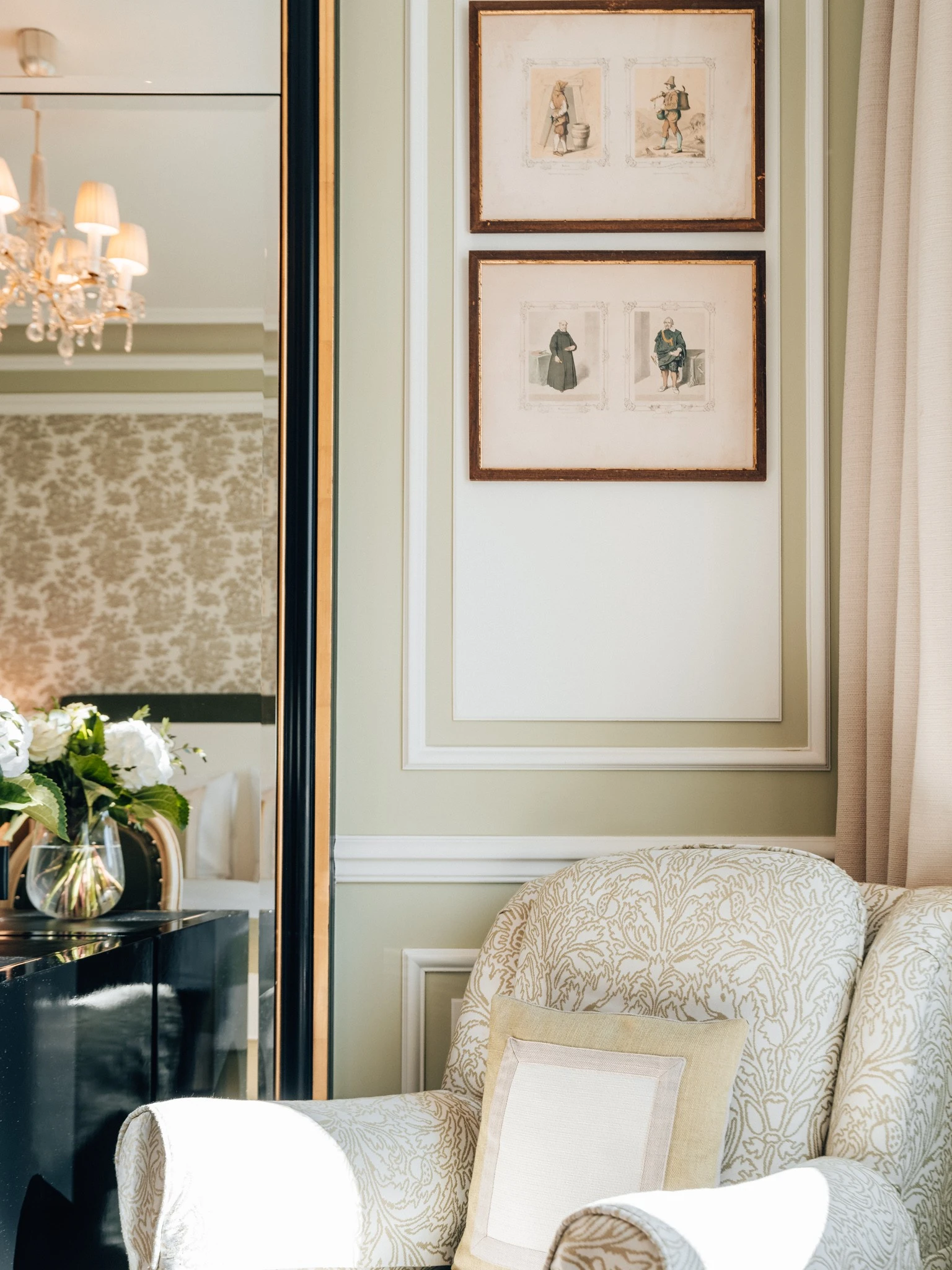 A cozy armchair with a patterned fabric and soft pillow sits beside a black desk with a vase of white flowers, beneath two framed vintage-style sketches on a wall bathed in natural light.
