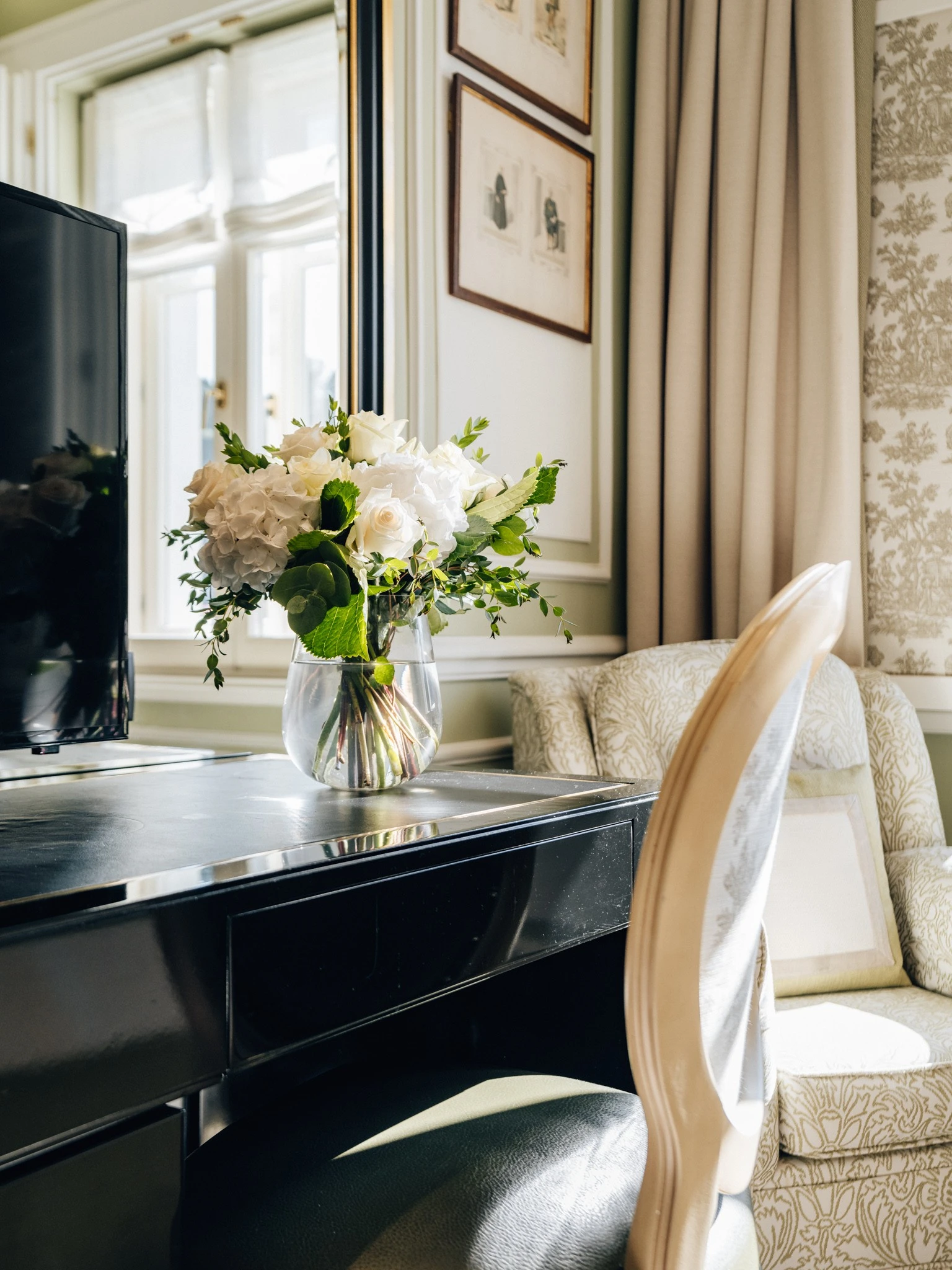 A glass vase filled with white roses and green leaves sits on a glossy black desk near a cushioned chair, with sunlight streaming through tall windows in a tastefully decorated room.