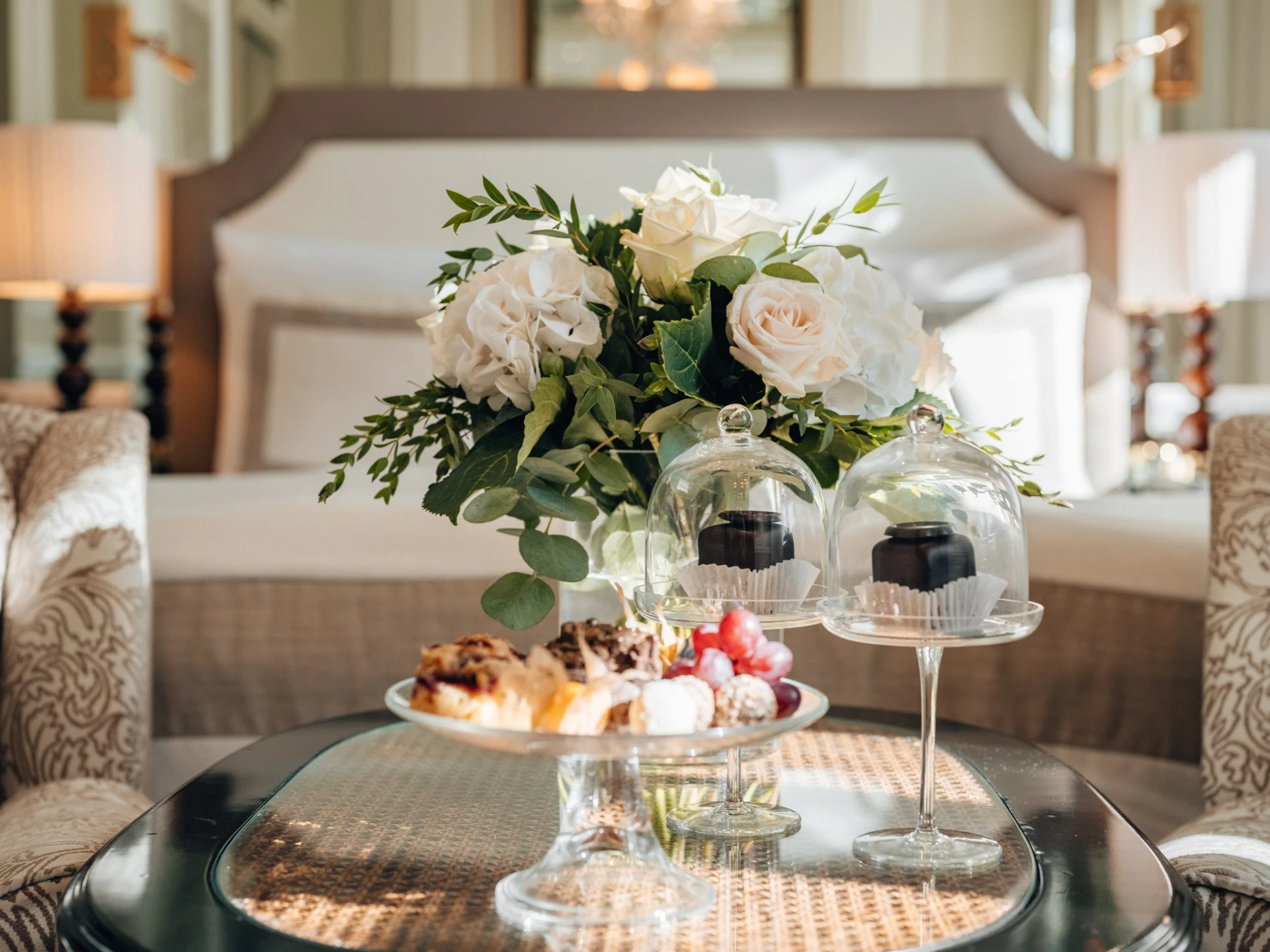 A luxurious bedroom features a small glass table topped with elegant desserts under glass domes, a plate of pastries and fruit, and a lush bouquet of white roses and greenery, all set against a plush bed in soft lighting.