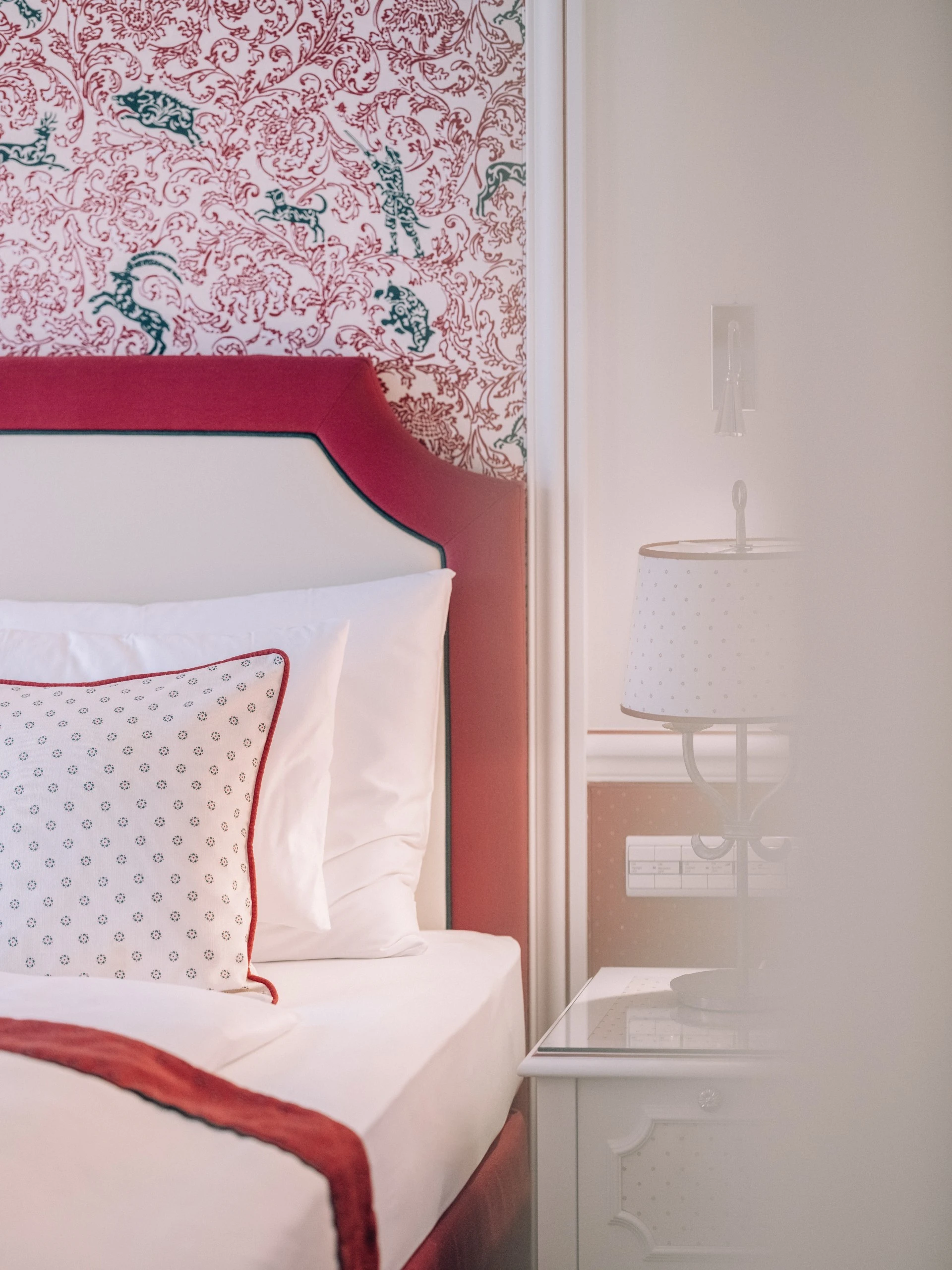 A cozy and elegant bedroom corner features a red and white color scheme with vintage floral wallpaper, a tufted headboard, patterned pillow, and a classic bedside lamp.