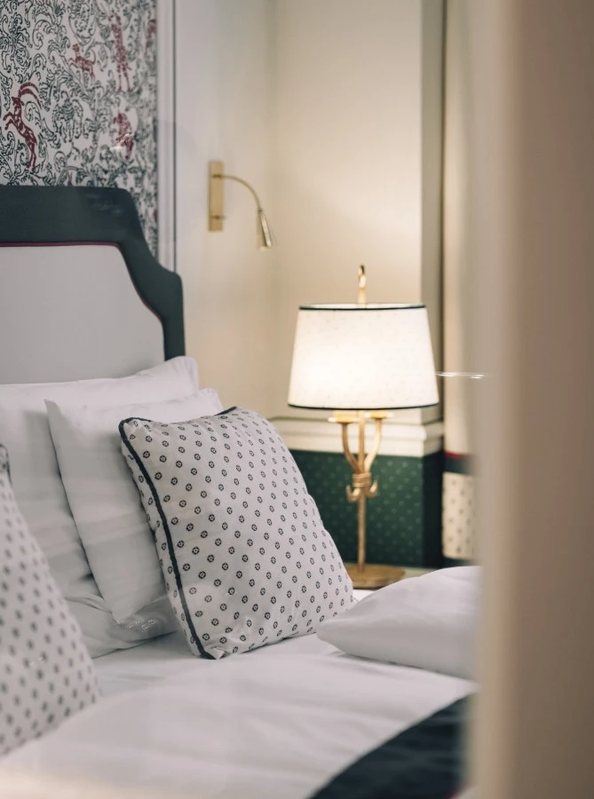 Junior-Suite-View_Hotel-Sacher-Salzburg_Print_28 (c) Hotel Sacher A close-up of an elegantly made bed with patterned pillows and a headboard, softly lit by a classic lamp and wall-mounted reading light, reflecting the refined charm of traditional decor.