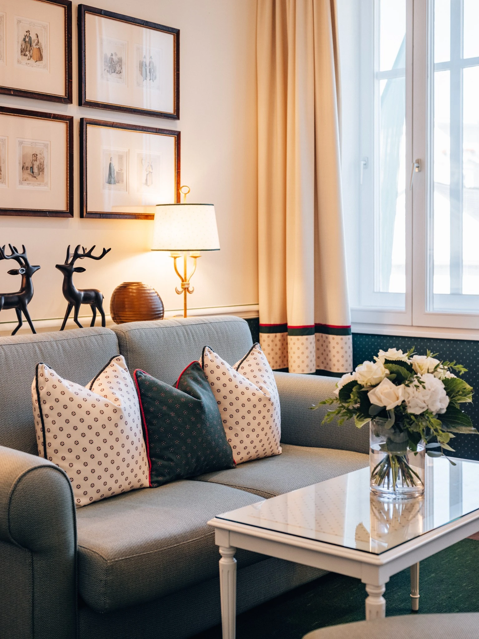 A cozy and tastefully decorated seating area featuring a soft blue-gray couch adorned with patterned pillows, elegant floral arrangements, and charming deer figurines, bathed in warm natural light from a nearby window.