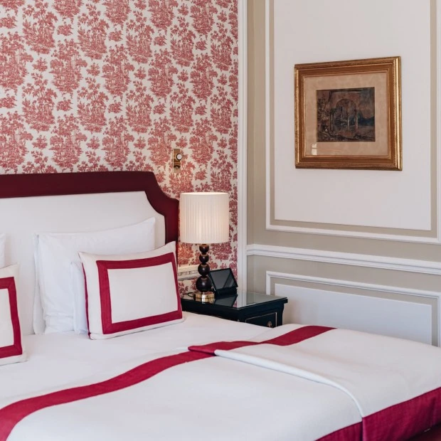 This photo showcases a luxurious hotel bed at Hotel Sacher Salzburg with crisp white linens accented by bold red borders, set against an ornate red and cream toile wallpaper, evoking a classic yet refined ambiance.