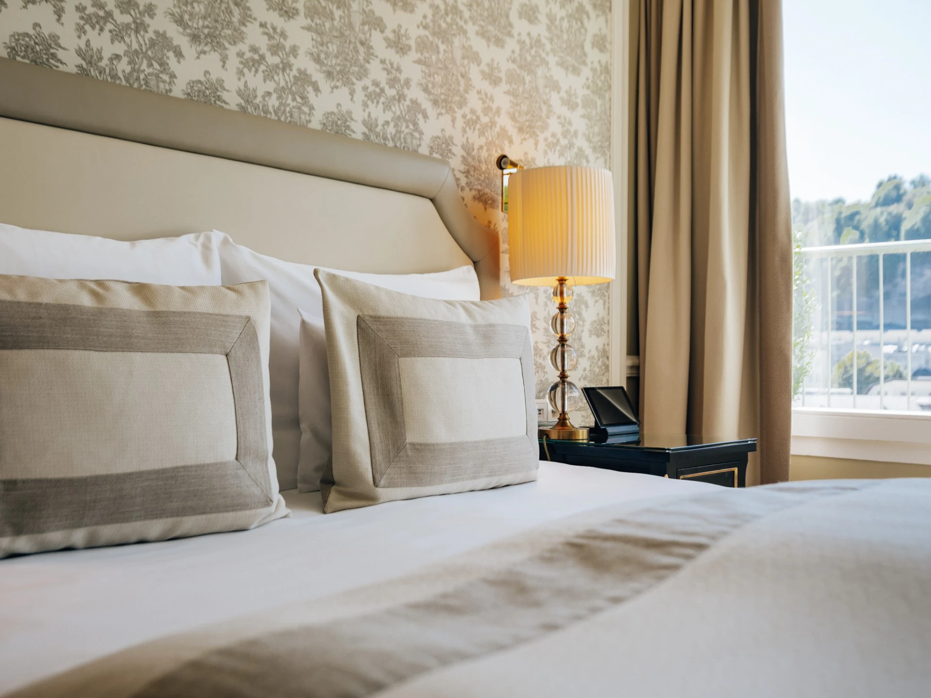 This image captures a serene and luxurious corner of a bedroom at Hotel Sacher Salzburg, with plush pillows, soft neutral bedding, elegant wallpaper, and natural light streaming through the window, offering a glimpse of the city beyond.