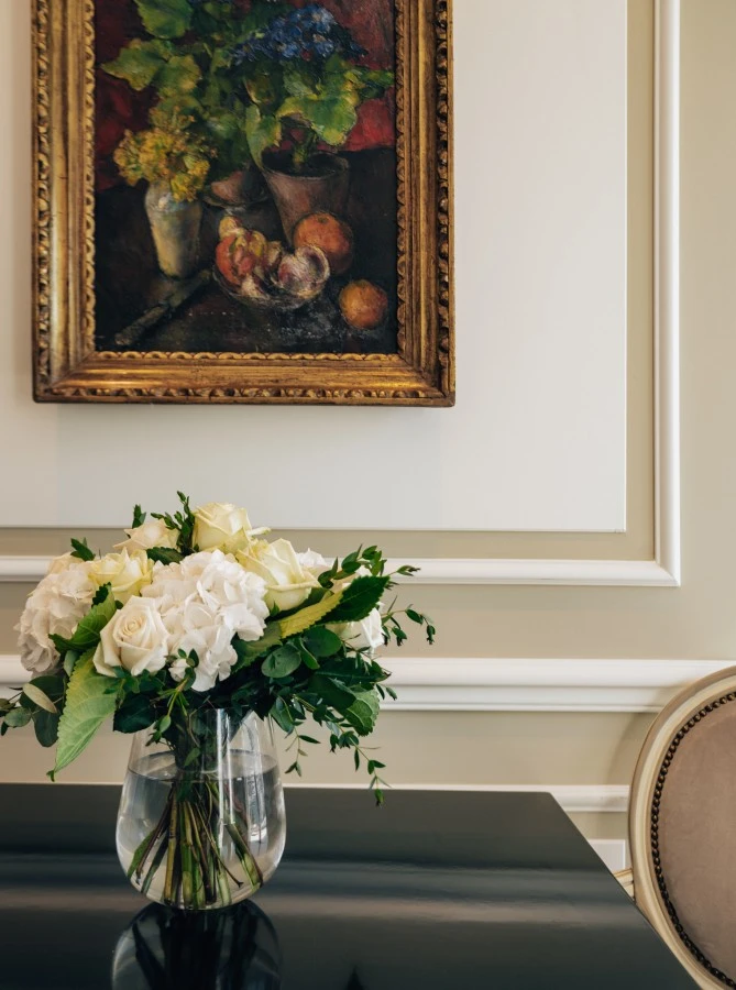 A lush bouquet of white roses and hydrangeas in a glass vase graces a glossy black tabletop, beneath a richly framed still-life painting at Hotel Sacher Salzburg.