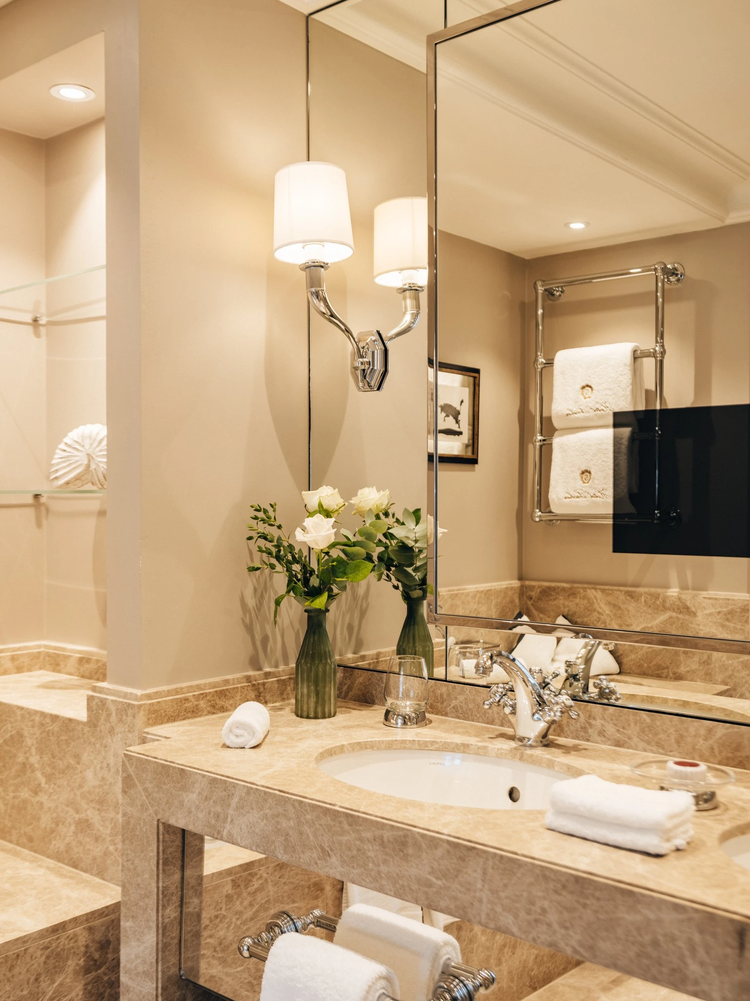 A luxurious marble bathroom at Hotel Sacher Salzburg features elegant lighting, fresh white roses, and neatly arranged towels, exuding timeless sophistication.
