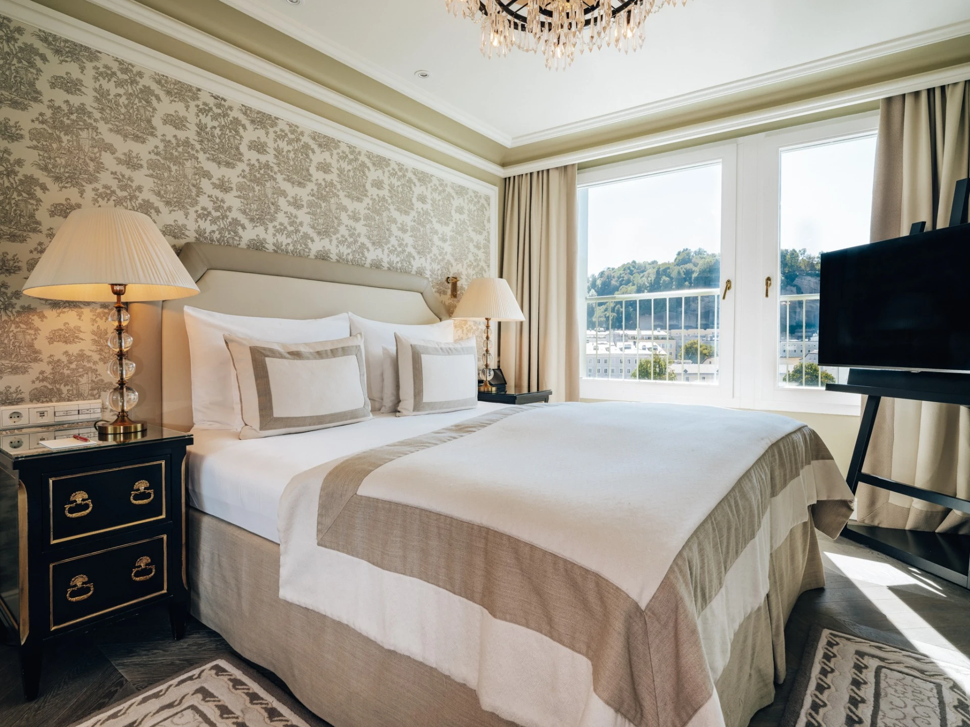 This serene bedroom at Hotel Sacher Salzburg combines elegant neutrals, classic wallpaper, and panoramic views of the city, offering refined comfort in a timeless setting.