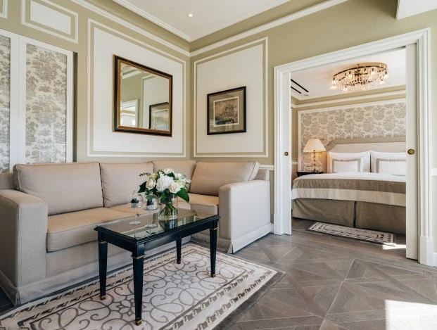 This elegant and spacious living area in the Presidential Suite at Hotel Sacher Salzburg features refined classic decor, plush seating, and a seamless view into the luxurious bedroom adorned with a statement chandelier.