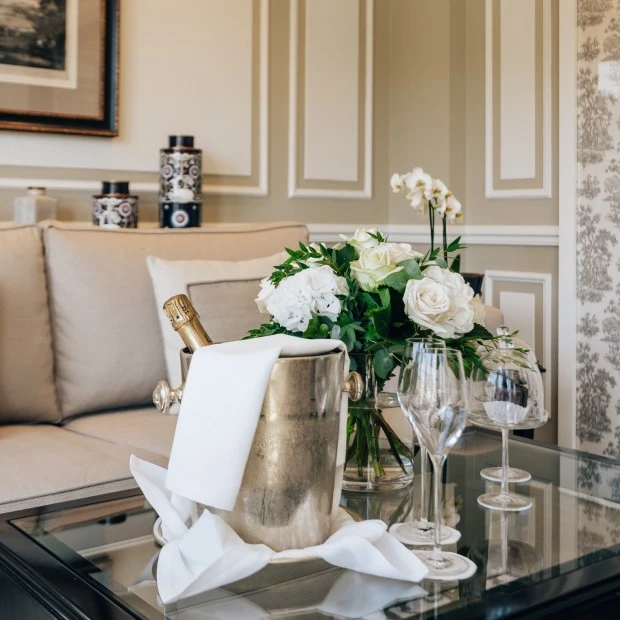 A refined corner at Hotel Sacher Salzburg showcases a glass table set with chilled champagne, crystal flutes, elegant white florals, and iconic Original Sacher Würfel, all framed by timeless interiors and Salzburg charm.