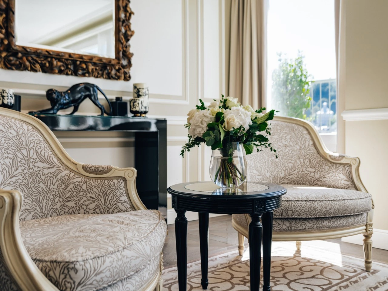 This elegant corner of Hotel Sacher features two classic armchairs flanking a small black table topped with fresh white flowers, all bathed in soft natural light from a nearby window.
