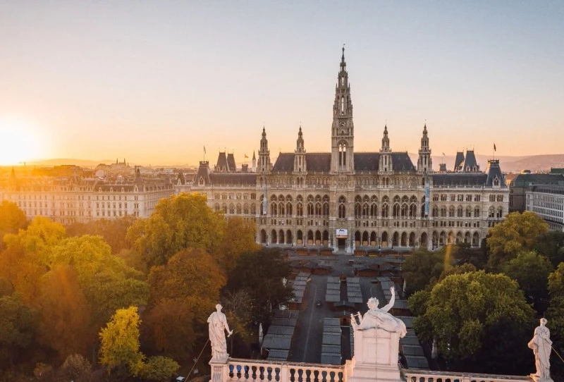 The Vienna City Hall (Rathaus) stands majestically at sunrise, surrounded by lush trees and classical statues, with soft golden light illuminating its neo-Gothic façade.