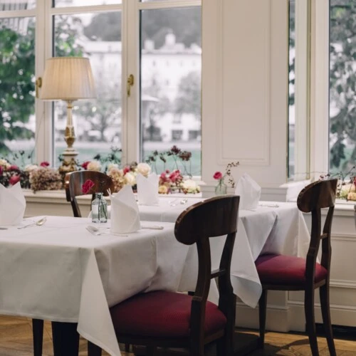 An elegant dining room with neatly set tables, burgundy-upholstered chairs, floral arrangements along the windowsill, and warm lighting from classic lamps, creating a cozy and refined atmosphere.
