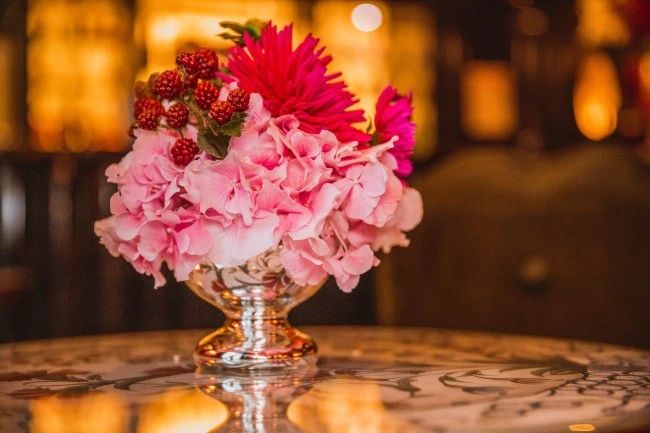 A vibrant bouquet of pink hydrangeas, red berries, and magenta blooms sits in an ornate silver vase on a reflective floral table, glowing warmly in the soft ambient light.