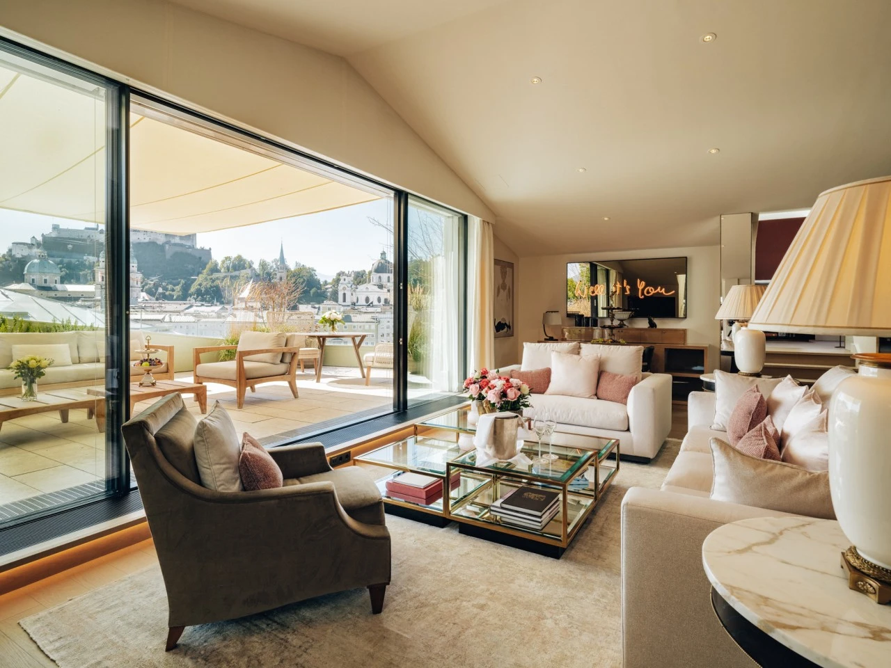 A bright and luxurious living space in the Sky Suite at Hotel Sacher Salzburg features soft pastel furnishings, fresh flowers, and floor-to-ceiling windows that open to a panoramic terrace with a stunning view of Salzburg’s historic skyline and Hohensalzburg Fortress.