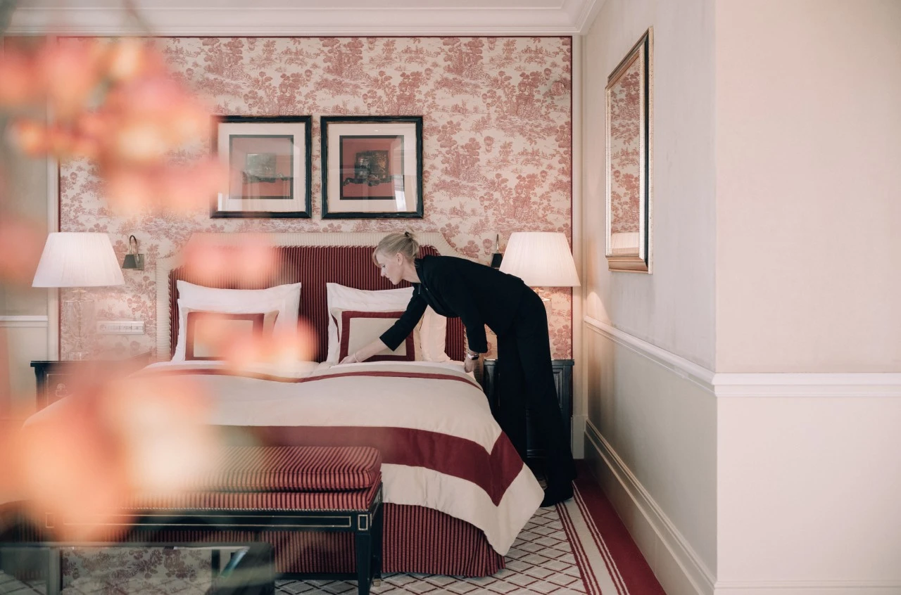 A staff member at Hotel Sacher Wien carefully perfects the bedding in a richly decorated room, where red and cream tones echo through the toile wallpaper, striped headboard, and matching bench, creating an atmosphere of refined luxury and meticulous attention to detail.