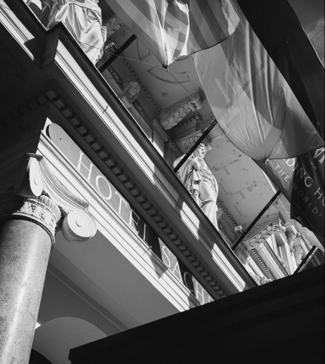 A dramatic black-and-white upward view of the grand entrance to Hotel Sacher Wien captures classical columns, intricate stonework, and statues beneath flowing flags, highlighting the building's timeless elegance and historic charm.
