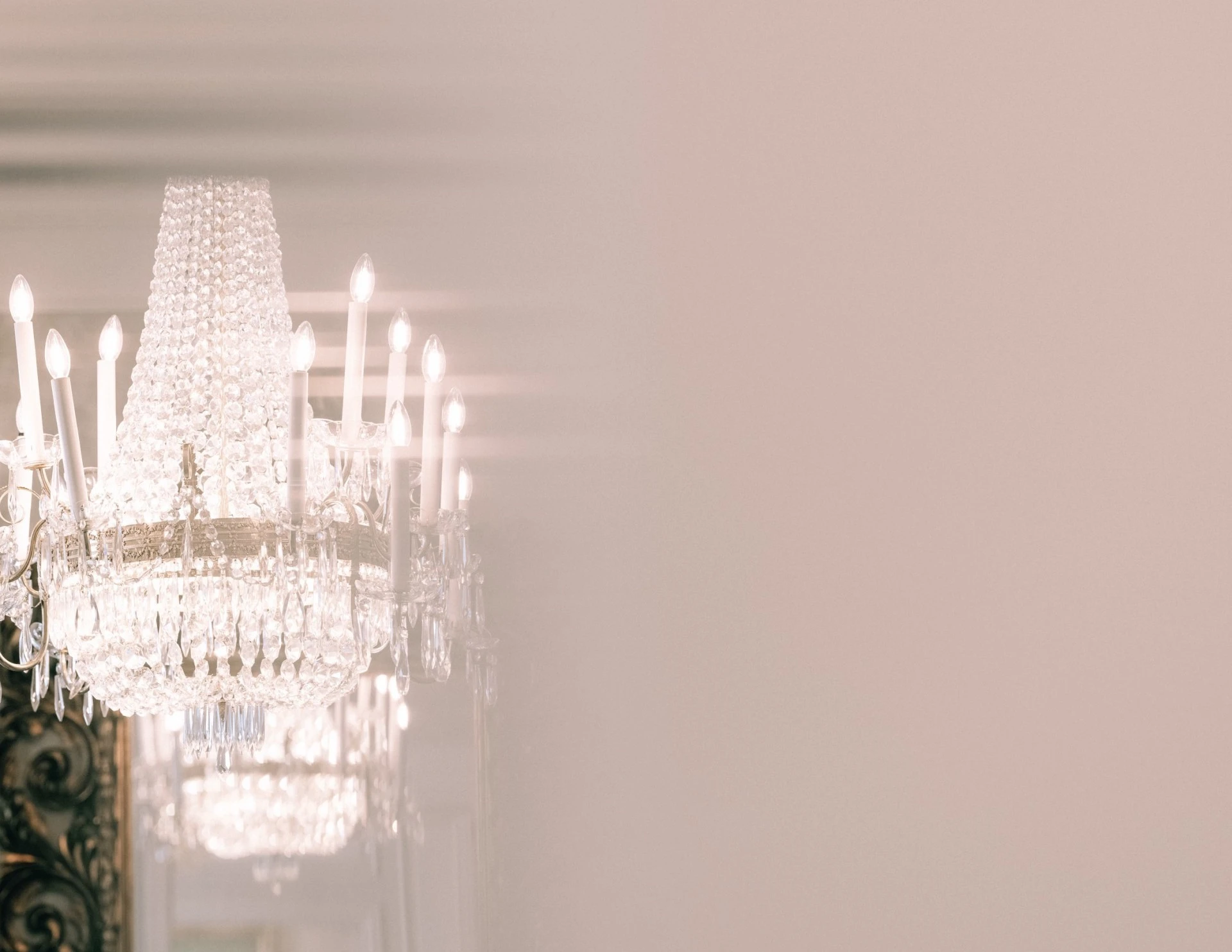 A grand crystal chandelier at Hotel Sacher Wien glows softly with candle-like bulbs, its delicate beads and facets casting elegant reflections in a mirror, partially obscured by a dreamy blur of soft pink light.