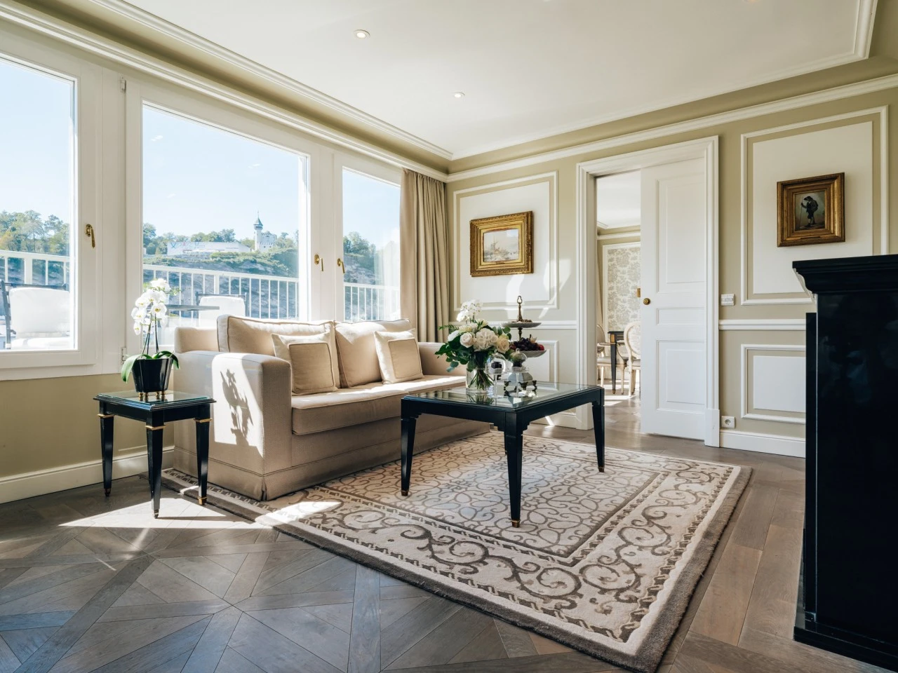 A sunlit suite at Hotel Sacher Salzburg features a cream-colored sofa, black lacquered furniture, fresh flowers, and large windows offering a sweeping view of the hills and fortress across the river, blending classic elegance with breathtaking scenery.