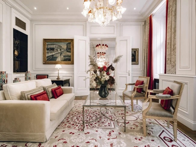 The Butterfly Suite at Hotel Sacher Wien radiates timeless elegance with cream-paneled walls, a sparkling crystal chandelier, antique-style armchairs, a plush sofa with red accent pillows, and a grand floral arrangement on a glass coffee table atop a richly patterned rug.