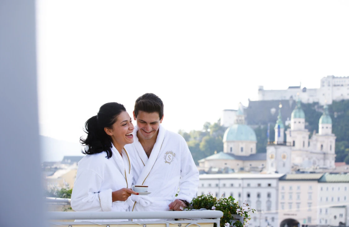 A smiling couple in white Hotel Sacher Salzburg robes enjoys a peaceful morning on a balcony, with a coffee cup in hand and the stunning historic skyline of Salzburg—including domed churches and the Hohensalzburg Fortress—rising in the background.