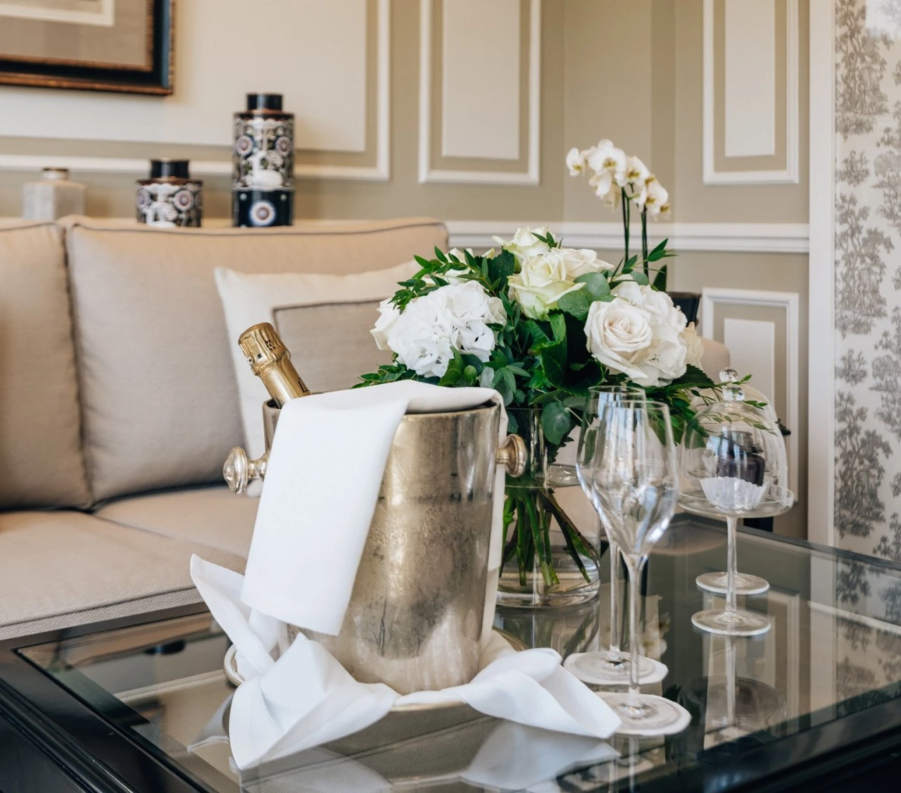 A romantic Valentine's Day setup at Hotel Sacher Salzburg features a champagne bucket with a bottle, crystal glasses, a bouquet of white roses, and a glass-covered dish of chocolates on a stylish glass table in a beautifully decorated suite.