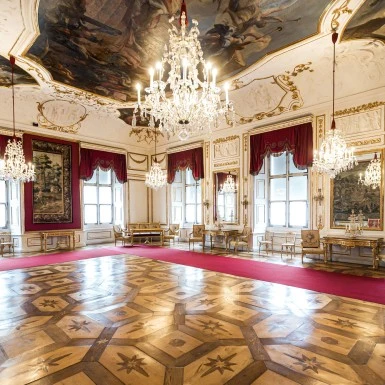 Prunkvoller Ballsaal mit kunstvollem Parkettboden, opulenten Kronleuchtern, reich verzierten Deckenmalereien, roten Samtvorhängen und vergoldeten Möbeln entlang der Wände.