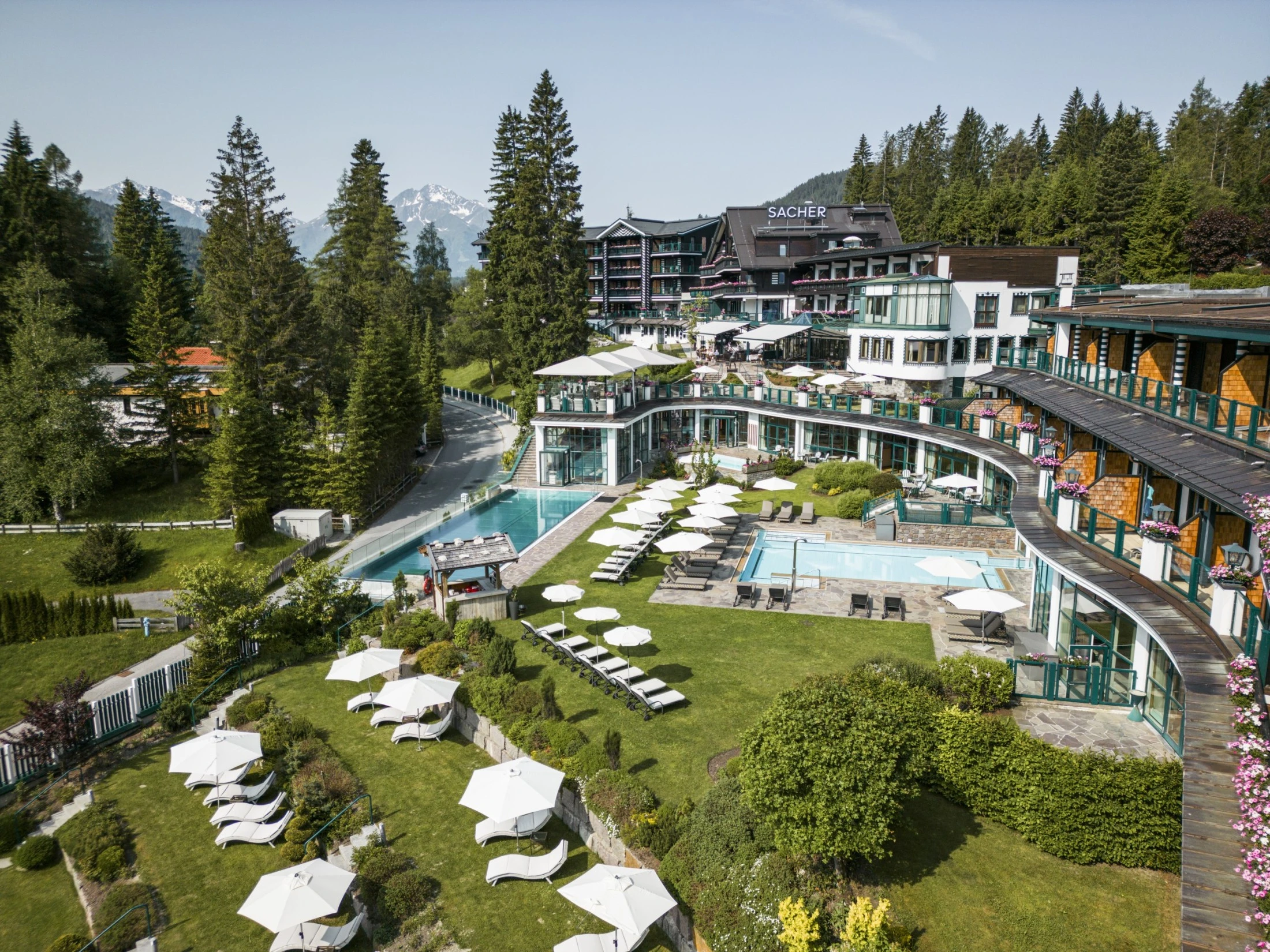 Aerial view of the Hotel Sacher Seefeld, showing its luxurious outdoor pool area, well-maintained gardens, sunbeds, and the surrounding mountains, with the hotel buildings in the background.