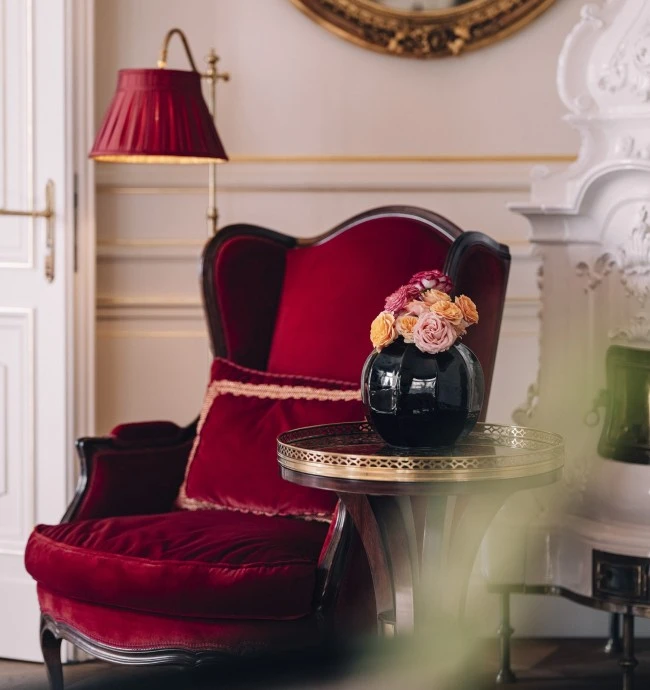 A refined and cozy corner of the Philharmonic Suite at Hotel Sacher Wien, featuring a red velvet armchair, a vase of colorful flowers, and a classic interior with a fireplace.
