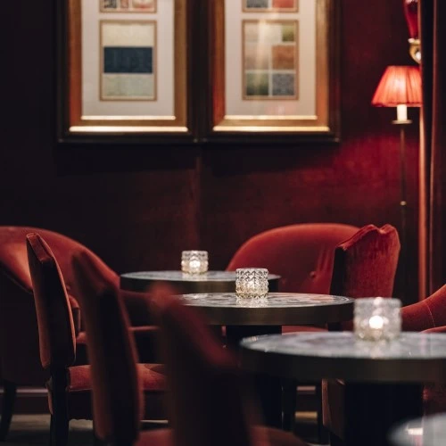 The intimate and stylish ambiance of the bar at Hotel Sacher Salzburg, with plush red velvet chairs and framed artwork on the walls.