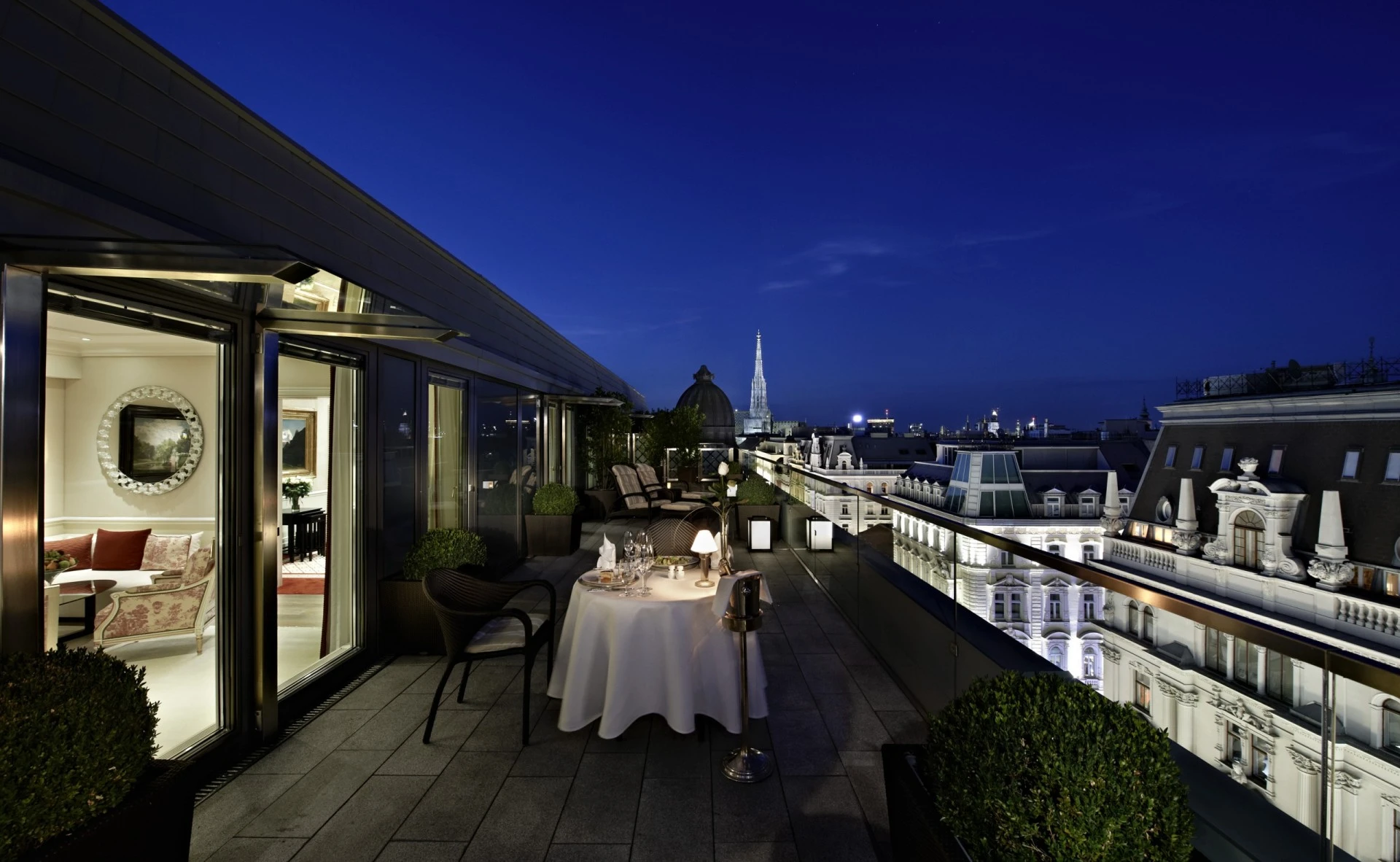 The breathtaking view from the Grand Signature Penthouse Suite at Hotel Sacher, featuring a private rooftop terrace with an intimate dining setup, offering a stunning view of Vienna's skyline at night.
