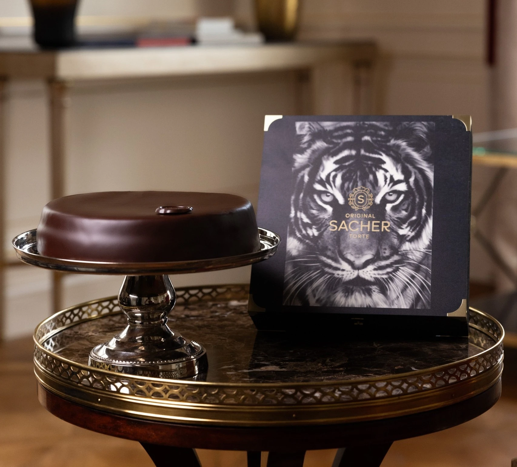 A beautifully presented Original Sacher Torte sits on a silver cake stand, accompanied by its limited edition box featuring a striking tiger illustration by Robert Longo, placed on a dark marble-topped table.