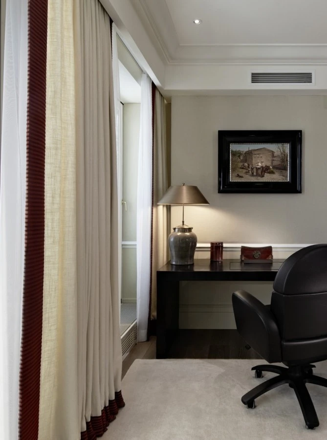 In the Grand Signature Penthouse Suite at Hotel Sacher, a sleek black writing desk with a modern lamp and elegant accessories sits beside tall, cream-colored curtains with burgundy accents, creating a refined and tranquil workspace.