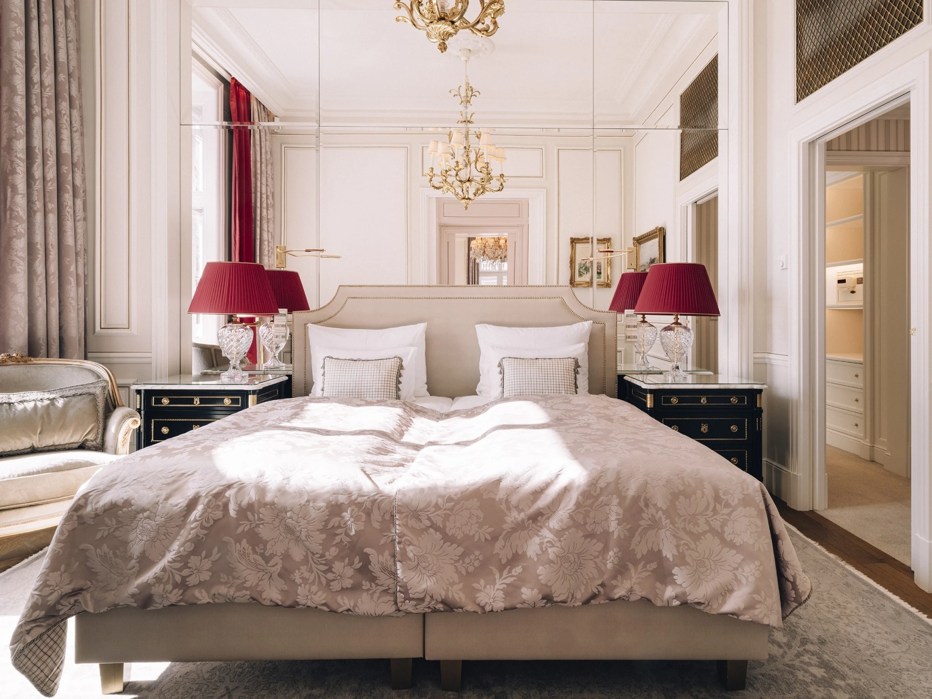 The bedroom of the Philharmonic Suite at Hotel Sacher features a grand bed with a floral-patterned duvet, flanked by black lacquered nightstands and crystal lamps with deep red shades, all bathed in natural light from tall windows framed by elegant drapes.