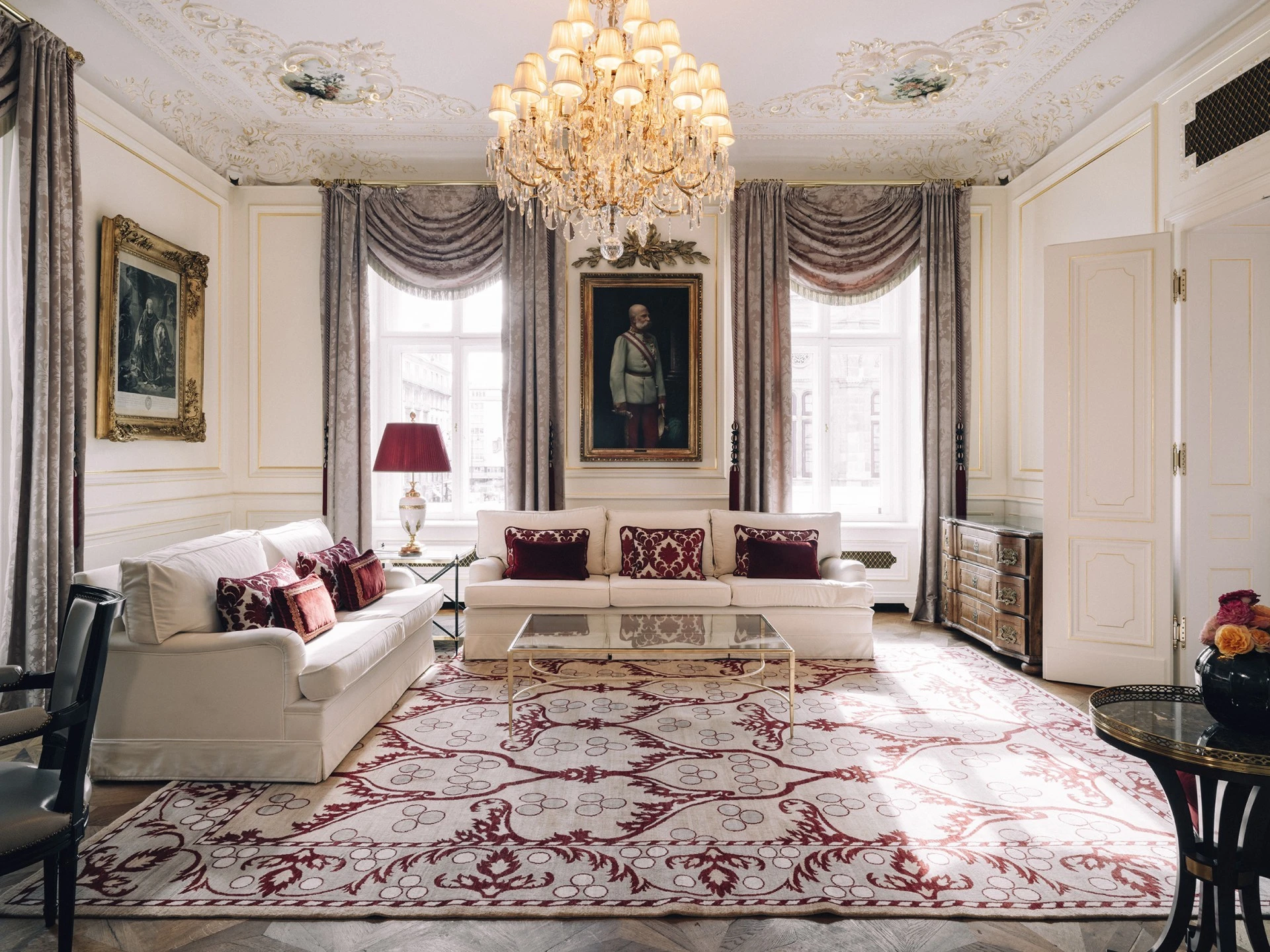 The Philharmonic Suite at Hotel Sacher exudes imperial elegance with ornate ceiling details, crystal chandeliers, and large windows draped in luxurious fabric, while white sofas with burgundy pillows sit atop a grand patterned rug, framing a portrait of Emperor Franz Joseph I.