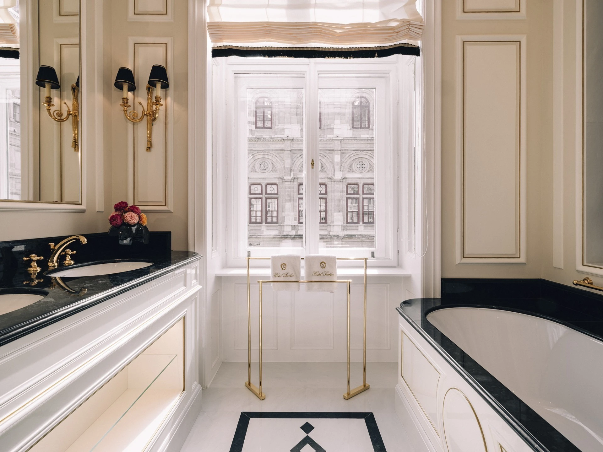 The bathroom of a Grand Signature Suite at Hotel Sacher features a luxurious black and white design with gold accents, a freestanding bathtub, double sinks with golden fixtures, and a window offering a stunning view of the Vienna State Opera.