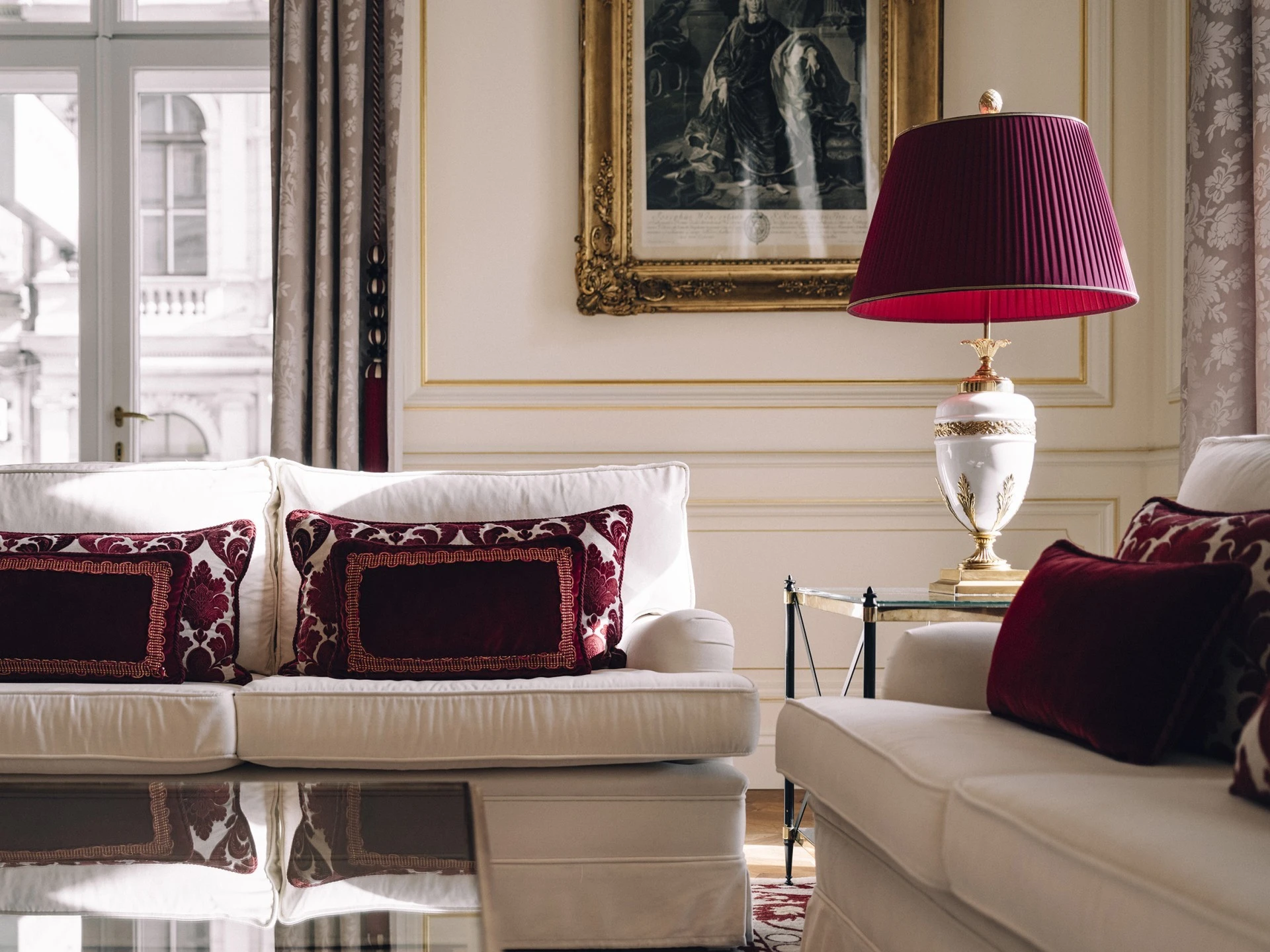 Inside a Grand Signature Suite at Hotel Sacher, elegant white sofas adorned with rich burgundy velvet pillows sit beneath a golden-framed artwork, with a regal white and gold table lamp topped by a pleated red shade adding a warm, luxurious touch.