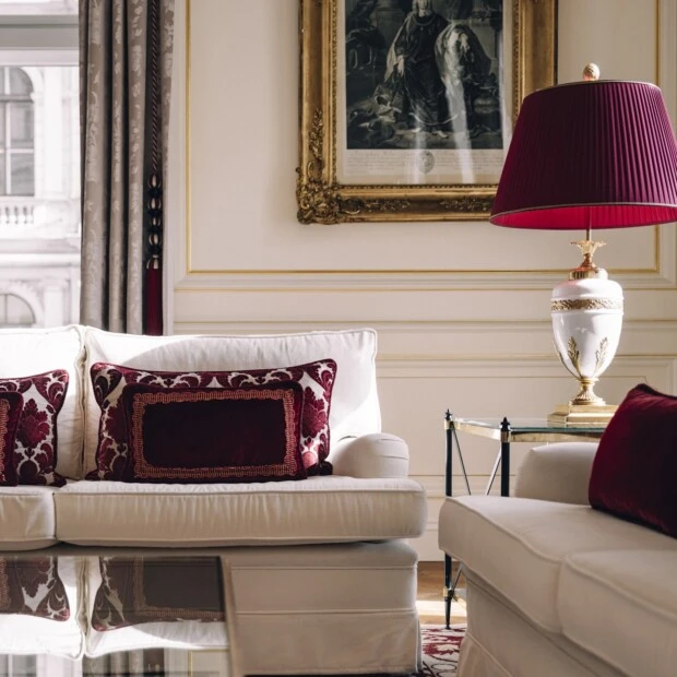 Inside a Grand Signature Suite at Hotel Sacher, elegant white sofas adorned with rich burgundy velvet pillows sit beneath a golden-framed artwork, with a regal white and gold table lamp topped by a pleated red shade adding a warm, luxurious touch.