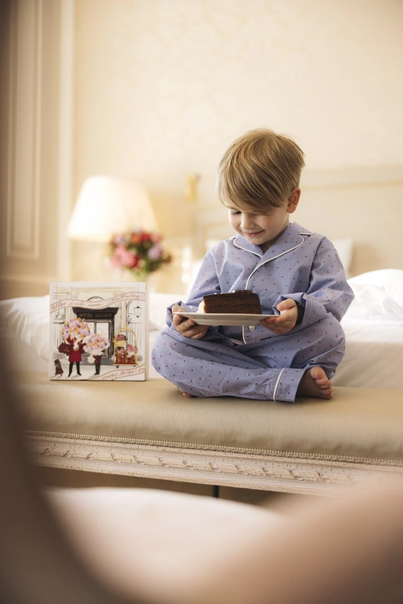 A young boy in light blue pajamas sits cross-legged on a bench in a cozy hotel room, smiling down at a plate holding a slice of Original Sacher Torte with whipped cream, beside a decorative Hotel Sacher gift box.
