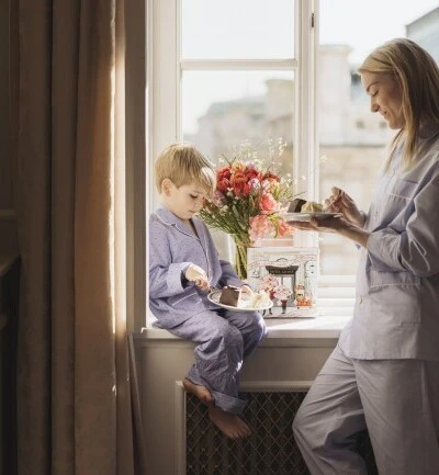 ChatGPT: In a cozy room at Hotel Sacher Vienna, a mother and her young son in pajamas enjoy slices of Sacher cake by a sunlit window, with fresh flowers and a decorative Original Sacher Torte box beside them.