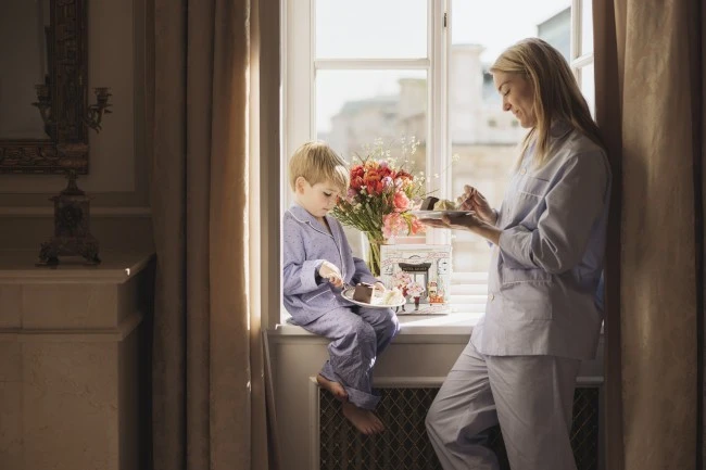 ChatGPT: In a cozy room at Hotel Sacher Vienna, a mother and her young son in pajamas enjoy slices of Sacher cake by a sunlit window, with fresh flowers and a decorative Original Sacher Torte box beside them.