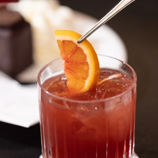 A close-up of a deep red Campari cocktail in a textured glass, being garnished with a fresh orange slice using silver tongs, with a blurred Original Sacher Würfel and whipped cream in the background.