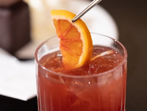 A close-up of a deep red Campari cocktail in a textured glass, being garnished with a fresh orange slice using silver tongs, with a blurred Original Sacher Würfel and whipped cream in the background.