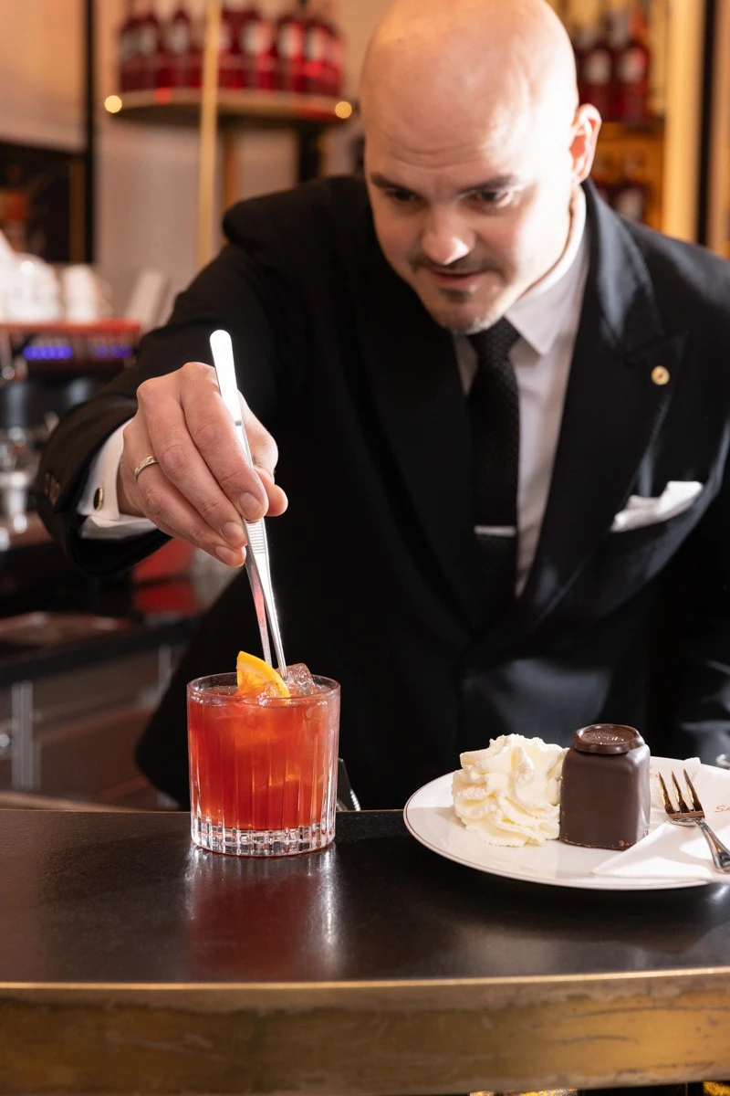 A bartender in a black suit carefully places an orange slice into a red Campari cocktail using silver tongs, next to a plate with an Original Sacher Würfel and a swirl of whipped cream.