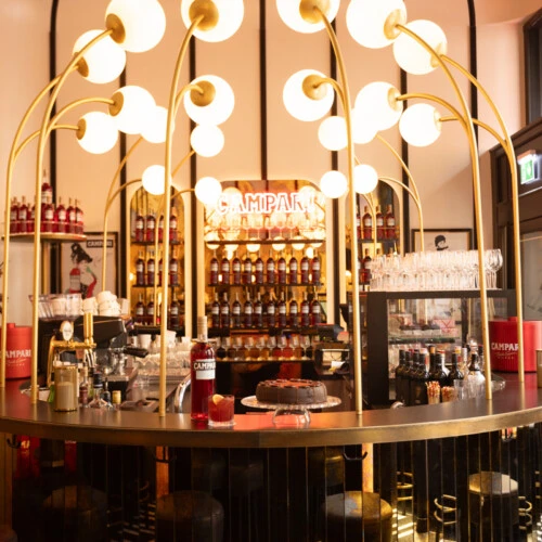 The bar at Salon Sacher is stylishly lit with sculptural golden light fixtures, showcasing a glowing Campari display, the Original Sacher-Torte, and elegant rows of bottles and glassware.