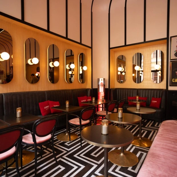 The interior of Salon Sacher features an elegant Campari-themed setup, with plush seating in pink and red tones, stylish black-and-white geometric flooring, and vintage Campari posters on the walls.