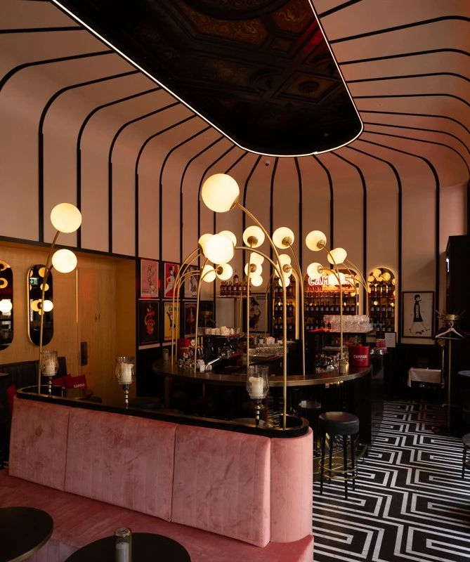 © Gregory Culbengan | www.adrianalmasan.com The bar area of Salon Sacher glows with warm, modern lighting under a striking arched ceiling design, featuring a sleek black counter, pink velvet seating, and walls adorned with vintage Campari posters.