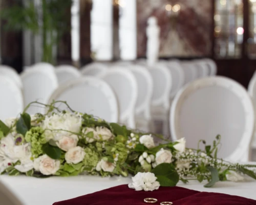 Close-up of two gold wedding rings on a red velvet pillow, placed next to a white floral arrangement in a ceremony room.