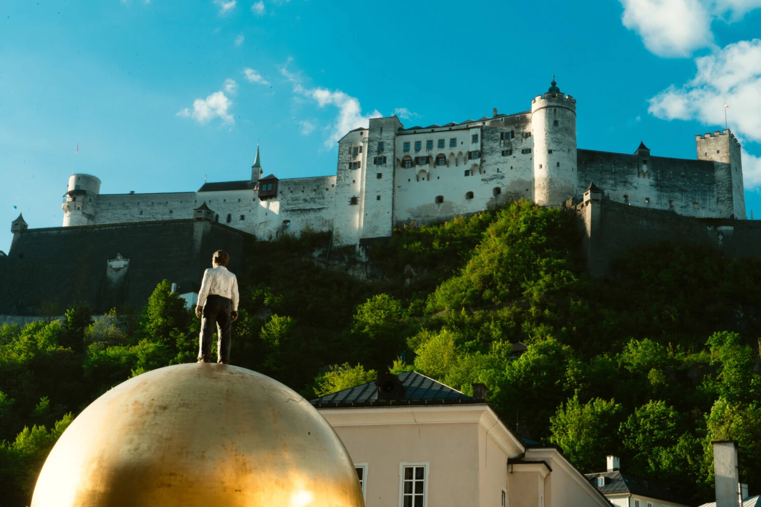 Fortress of Hohensalzburg surrounded by trees together with the statue of a child standing on a giant golden globe