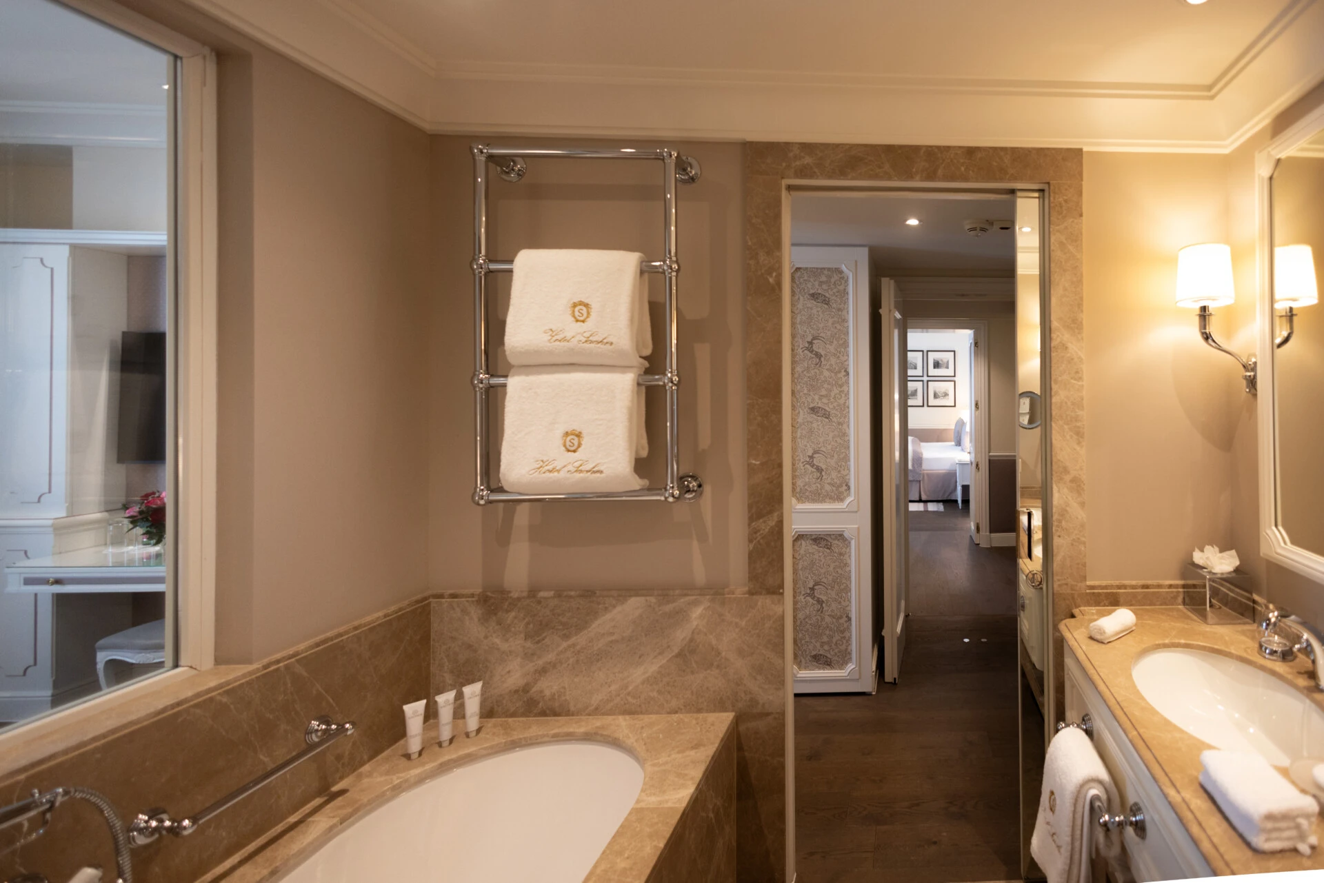 A warmly lit bathroom at Hotel Sacher Salzburg features a marble bathtub, a chrome towel warmer with folded white towels, a double sink vanity with marble countertops, and a glimpse into an adjacent room with a bed.